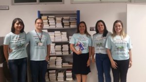 Equipe do CEAF (quatro pessoas, com camisetas da campanha) diante de uma estante cheia de livros.