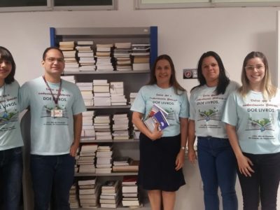 Equipe do CEAF (quatro pessoas, com camisetas da campanha) diante de uma estante cheia de livros.