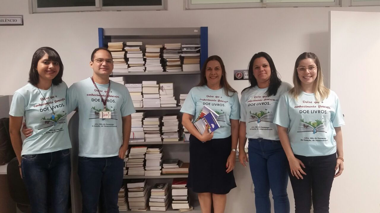 Equipe do CEAF (quatro pessoas, com camisetas da campanha) diante de uma estante cheia de livros.
