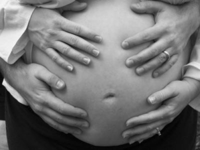 Barriga de gestante. Sobre ela, repousam as mãos da própria mulher e as mãos de um homem.