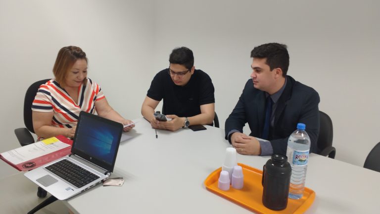 Três pessoas assinam documentos sentadas em torno de uma mesa