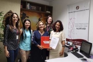 Mulheres de pé sorrindo para fotografia. Uma das mulheres segura uma pasta nas mãos com a marca do Ministério Público do Estado