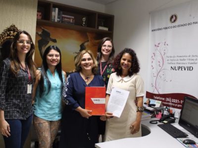 Mulheres de pé sorrindo para fotografia. Uma das mulheres segura uma pasta nas mãos com a marca do Ministério Público do Estado