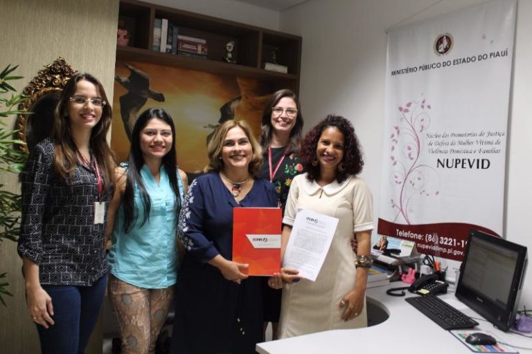 Mulheres de pé sorrindo para fotografia. Uma das mulheres segura uma pasta nas mãos com a marca do Ministério Público do Estado