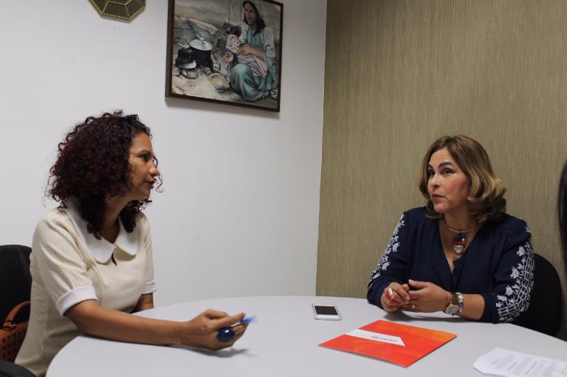Duas mulheres sentadas em volta de uma mesa, conversando. Sobre a mesa existe uma pasta com o brasão do Ministério Público 
