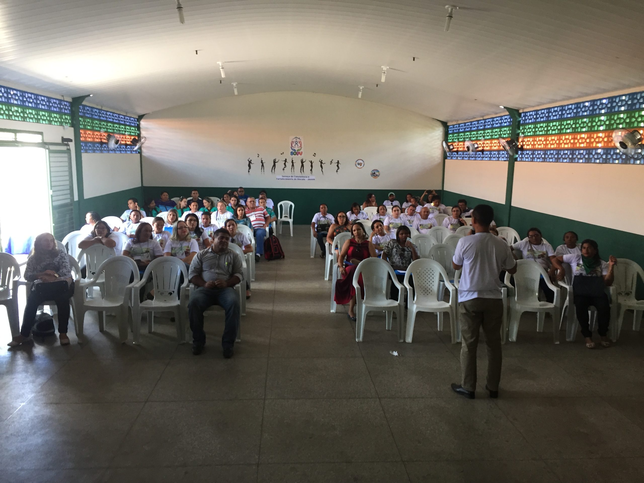Pessoas sentadas em um ginásio observando a palestra que está sendo proferida 
