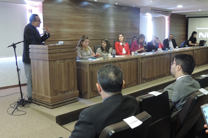 Homem de pé atrás de uma tribunal, falando ao micrfone ao mesmo tempo em que olha para mulheres sentadas ao lado 