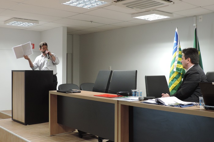 Homem de pé segurando uma pasta com uma das mãos e na outra um microfone 