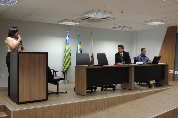 Mulher de pé com microfone na mão, dois homens sentados 