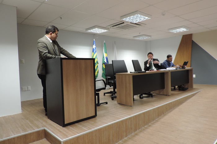 Homem de pé olhando para outro que está sentando falando com um microfone nas mãos 