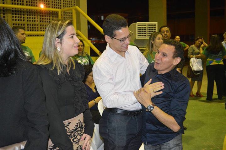 Três pessoas de pé, duas das três pessoas se abraçam e sorriem 