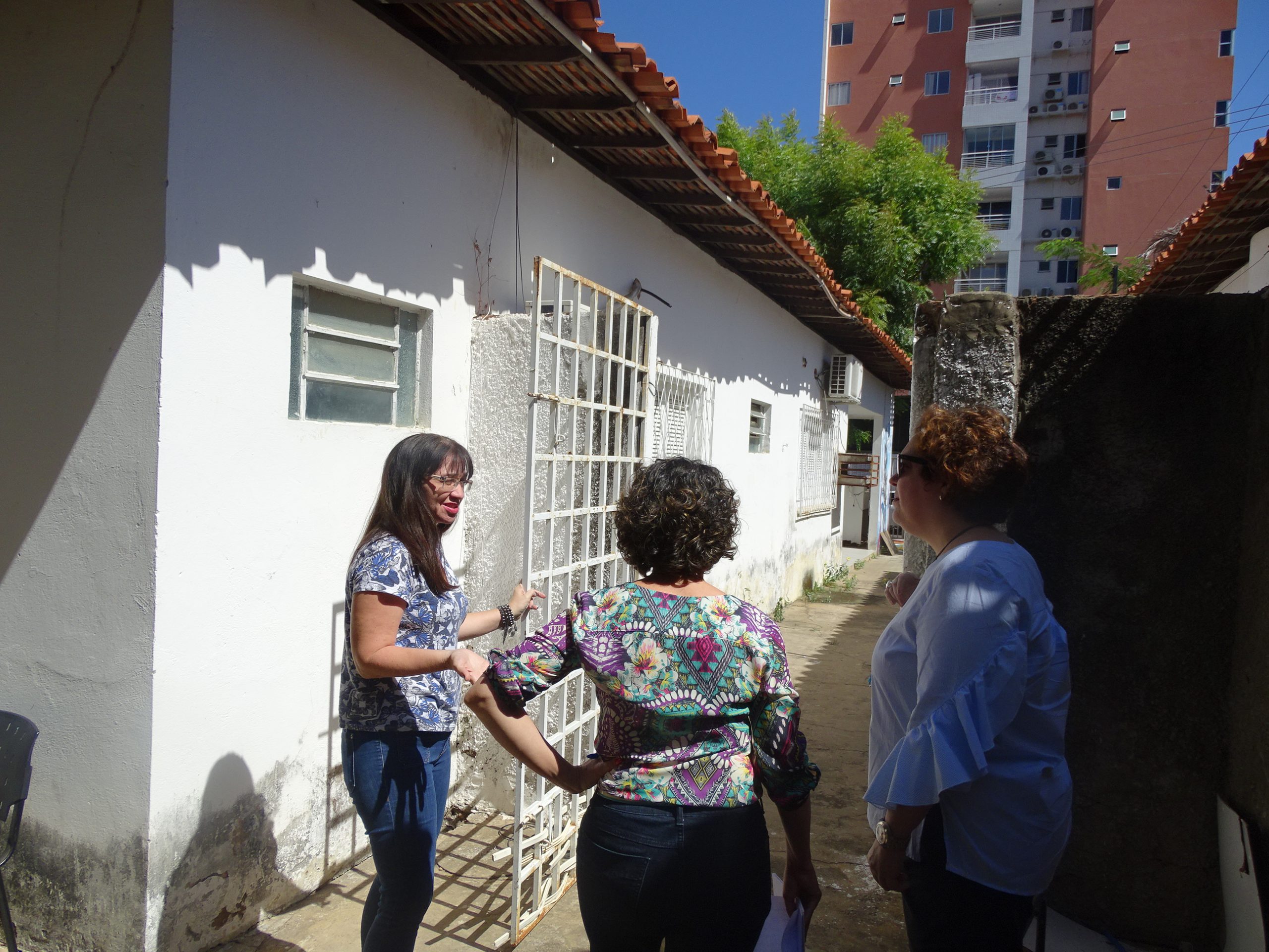 Três pessoas de pé na área externa uma uma casa. Uma das pessoas abre um portão enquanto conversa com as demais. 