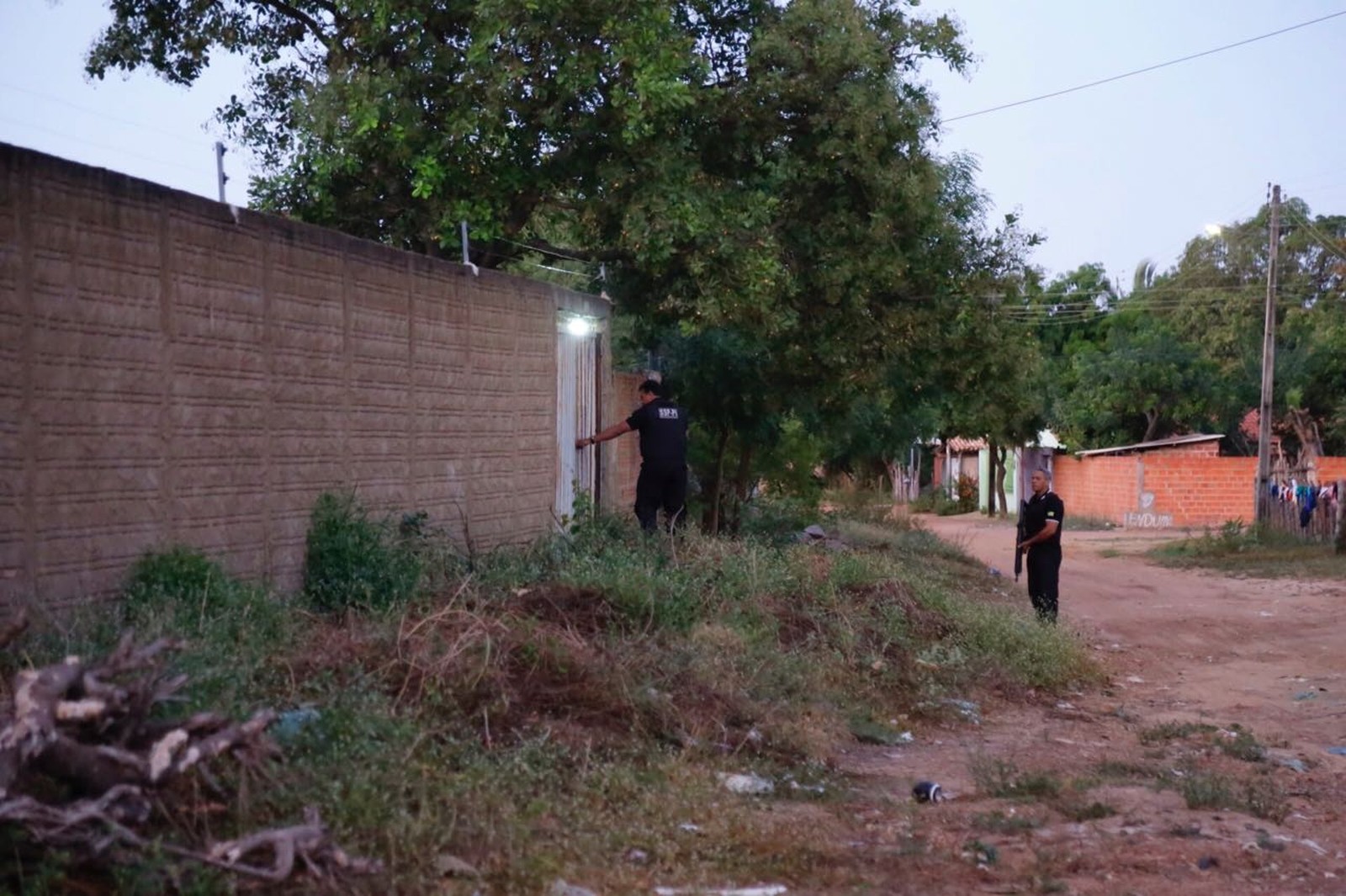 Duas pessoas de pé no meio de uma rua. Uma das pessoas está encostada no portão de uma casa 