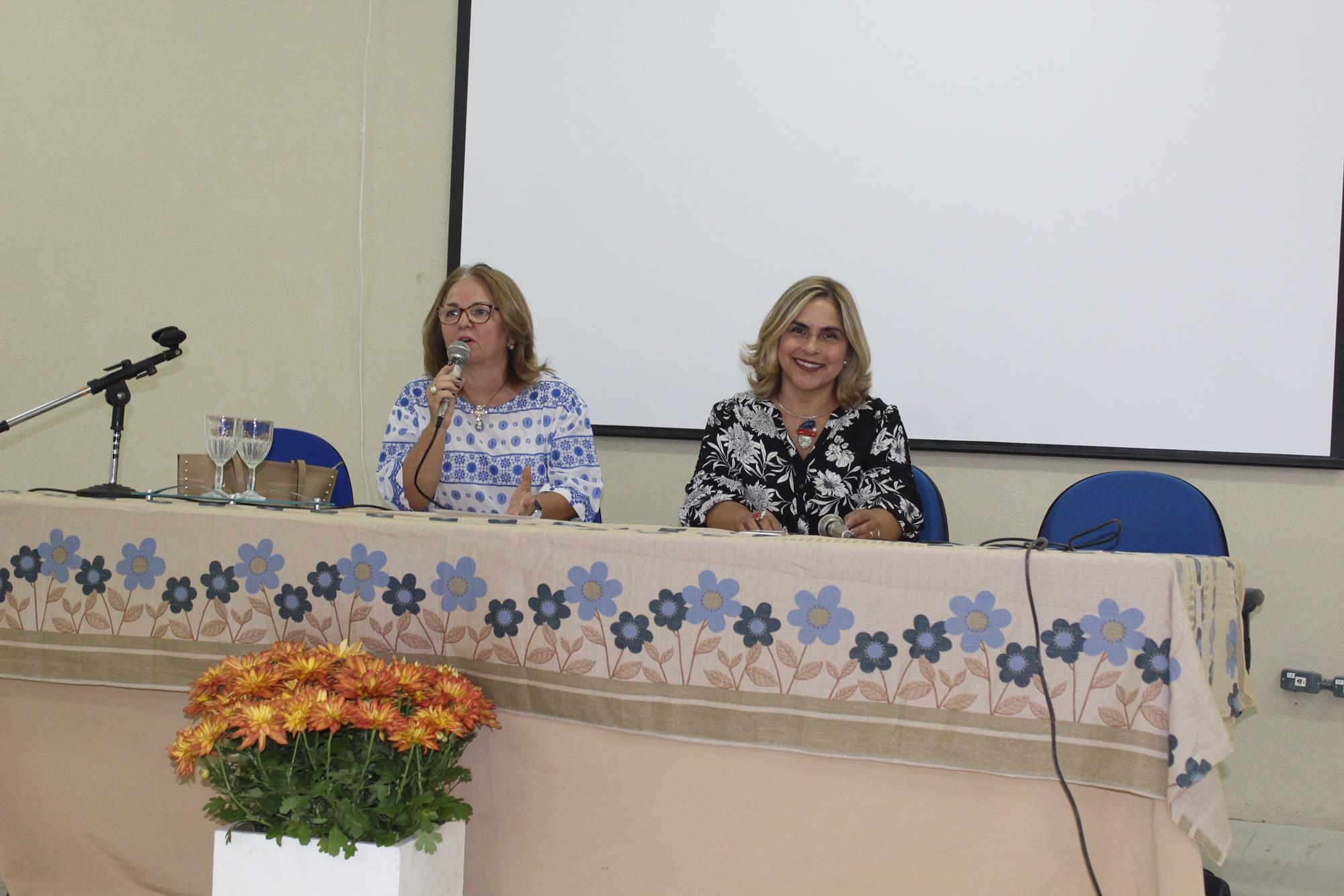 Duas pessoas sentadas em uma mesa. Uma das pessoas segura o microfone nas mãos e fala 