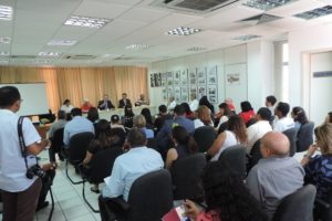 Pessoas sentadas em audiência. Ao fundo está uma mesa com pessoas sentadas