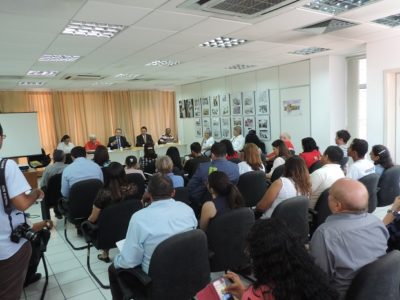 Pessoas sentadas em audiência. Ao fundo está uma mesa com pessoas sentadas