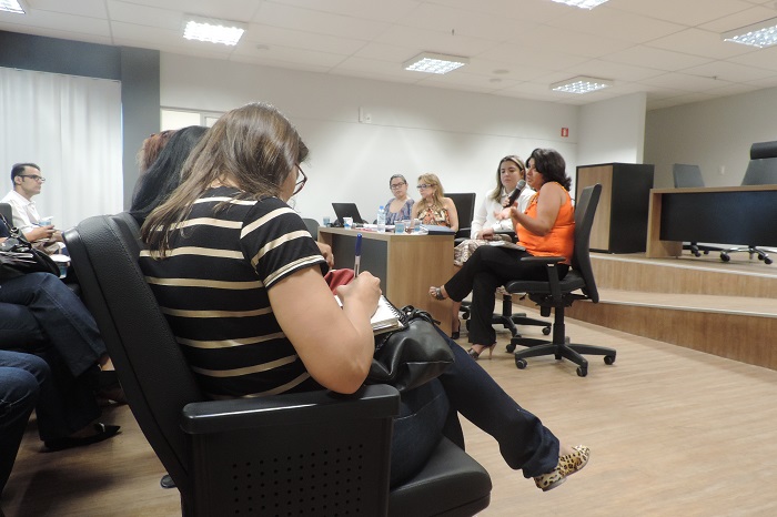 Pessoa sentada com caderno nas mãos enquanto anota há uma pessoa ao fundo com microfone nas mãos 