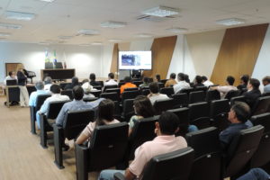 Pesoas sentadas em auditório olhando para uma televisão ao fundo que exibia matéria sobre o tema da reunião