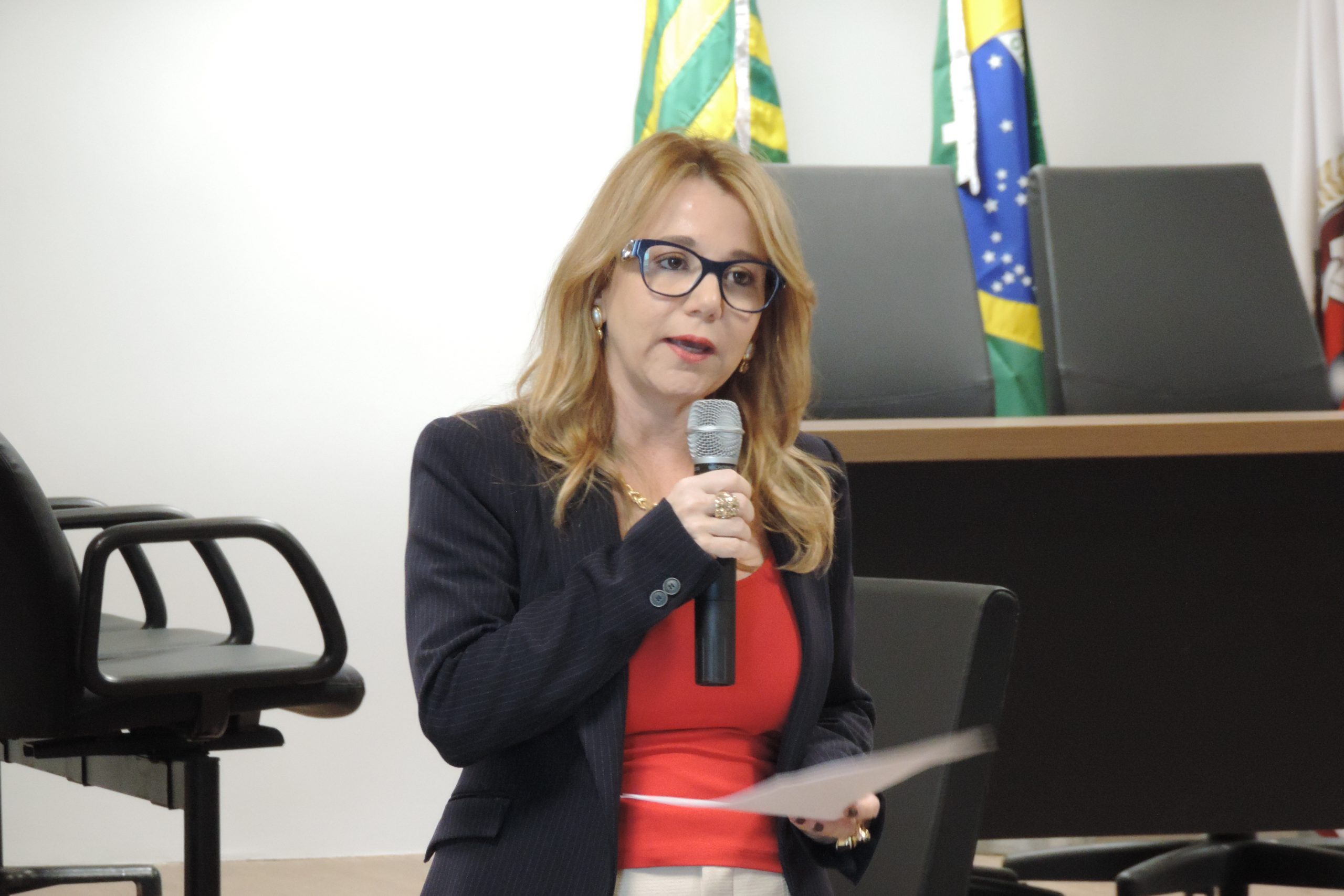 Mulher de pé. numa das mãos segura um microfone, na outra balança uma folha de papel 