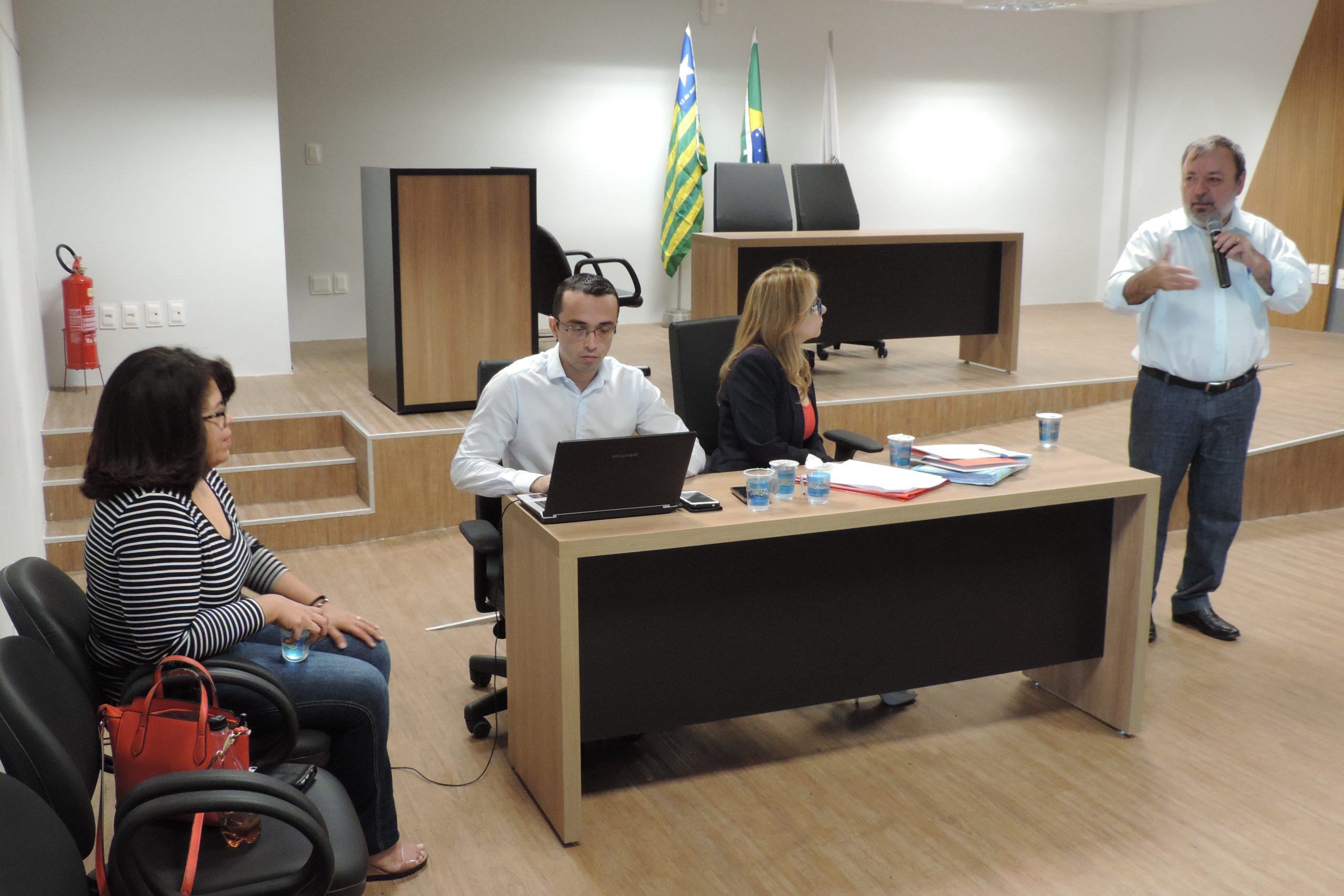 Quatro pessoas, três estão sentadas, duas observam o homem que está de pé falando com microfone nas mãos. A outra pessoas está sentadas escrevendo no computador