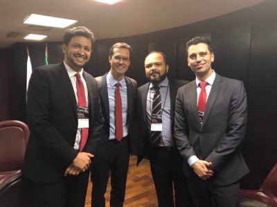 Quatro homens em pé de terno, em frente a banner de evento