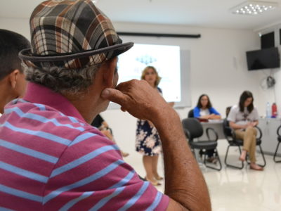 Sala com vários homens sentados. A Promotora de Justiça fala a eles, de pé.