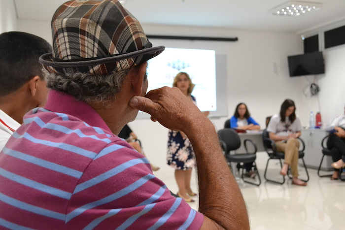 Sala com vários homens sentados. A Promotora de Justiça fala a eles, de pé.