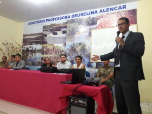 Promotor de Justiça fala, de pé, diante de uma mesa de honra