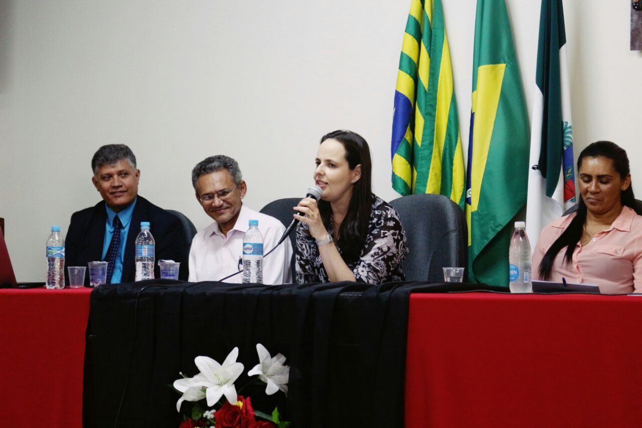 Promotora de Justiça fala ao microfone em mesa de honra