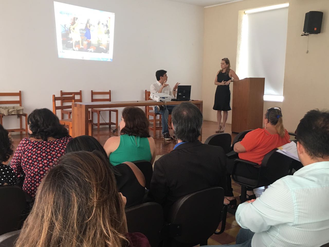 Pessoas sentadas em auditório olhando para outras duas pessoas que estão mais a frente falando sobre o projeto  