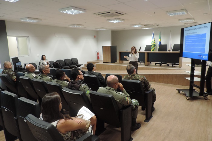 Pessoas sentadas em auditório quanto mulher está de pé falando ao microfone 