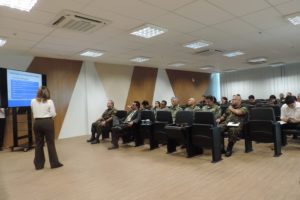 Pessoas sentadas em auditório. Uma mulher está de pé com microfone nas mãos
