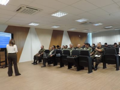 Pessoas sentadas em auditório. Uma mulher está de pé com microfone nas mãos