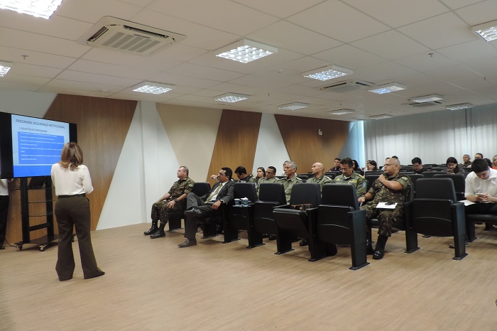 Pessoas sentadas em auditório. Uma mulher está de pé com microfone nas mãos