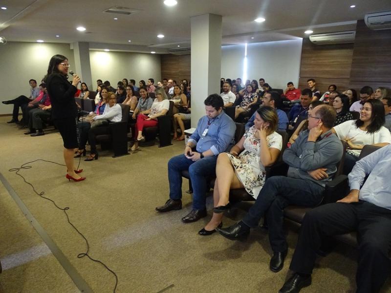 Pessoas sentadas observando uma mulher que está de pé falando com microfone nas mãos 