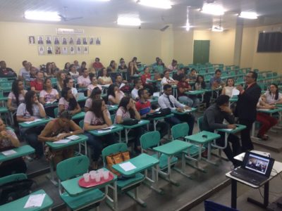 Pessoas sentadas em auditório, a frente das pessoas tem um homem de pé com microfone nas mãos