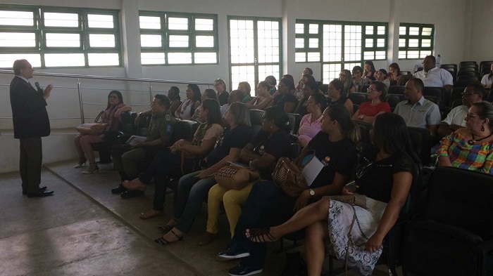 Pessoas sentadas em auditório. A frente dessas pessoas está um homem de pé segurando um microfone com uma das mãos. 