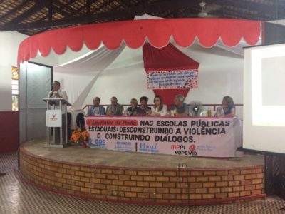 Palco de um local fechado. De pé há um homem em uma tribuna que discusa para as pessoas que o assistem. Um pouco mais ao fundo existe uma mesa com pessoas sentadas e um cartaz na frente da mesa