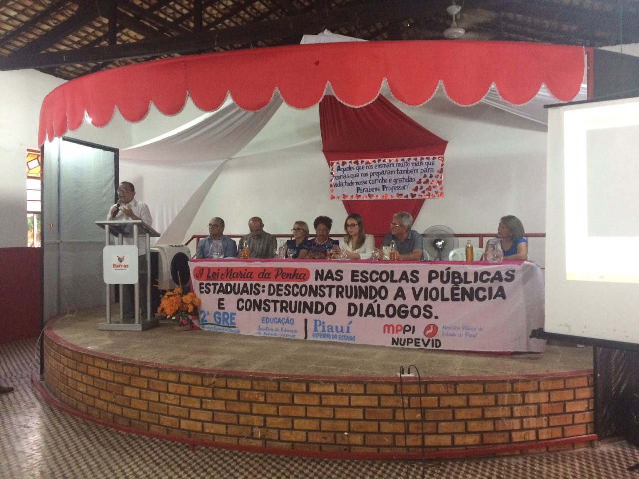 Palco de um local fechado. De pé há um homem em uma tribuna que discusa para as pessoas que o assistem. Um pouco mais ao fundo existe uma mesa com pessoas sentadas e um cartaz na frente da mesa 