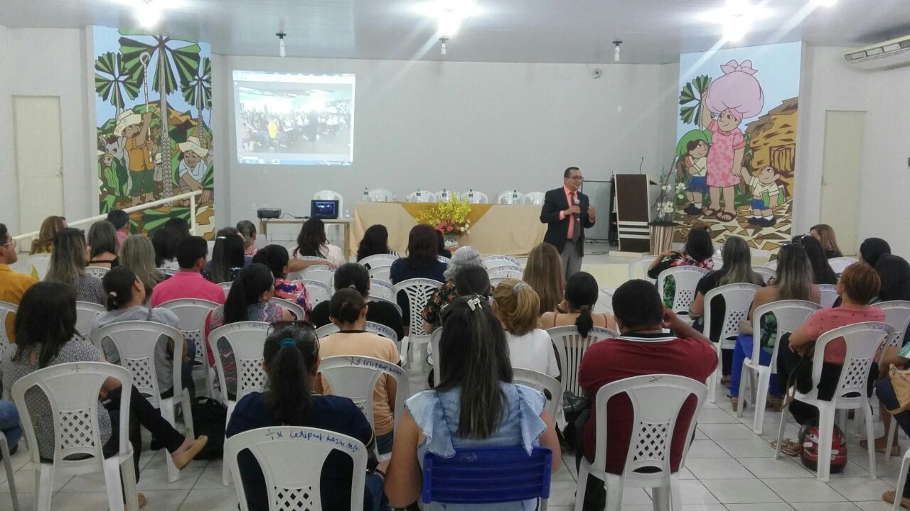 Pessoas sentadas em uma sala observam ao fundo um homem de fala que fala segurando um microfone numa das mãos