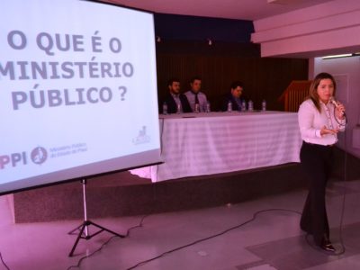 Mulher de pé se movimentando com microfone nas mãos enquanto fala para platéia que está na sua frente