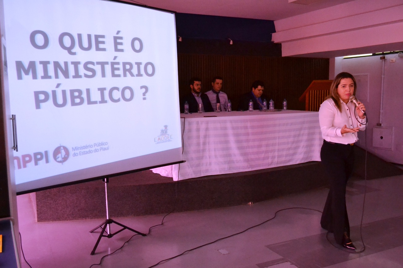 Mulher de pé se movimentando com microfone nas mãos enquanto fala para platéia que está na sua frente