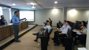Pessoas sentadas em uma auditório. Logo a frente dessas pessoas um home está de falando e gesticulando com as mãos, mais ao fundo uma pessoa está assentada em uma bancada
