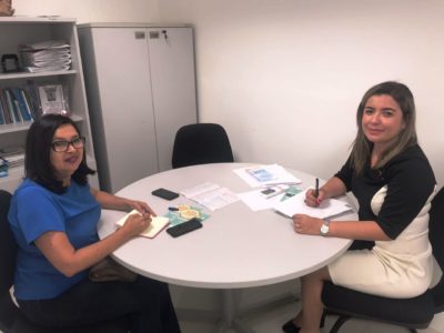 Duas pessoas sentadas em volta de uma mesa com vários papéis sobre a mesma. No momento as duas pessoas estão olhando para a pessoa que faz o registro fotográfico da reunião