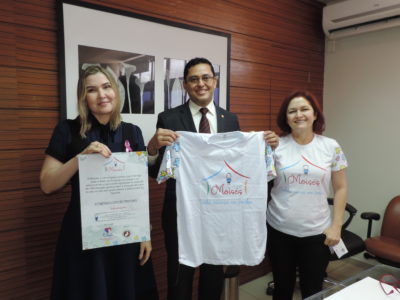 Três pessoas de pé. Uma segura um cartaz, outra uma camisa e a última pessoa segura um pequeno folder informativo