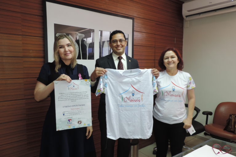 Três pessoas de pé. Uma segura um cartaz, outra uma camisa e a última pessoa segura um pequeno folder informativo