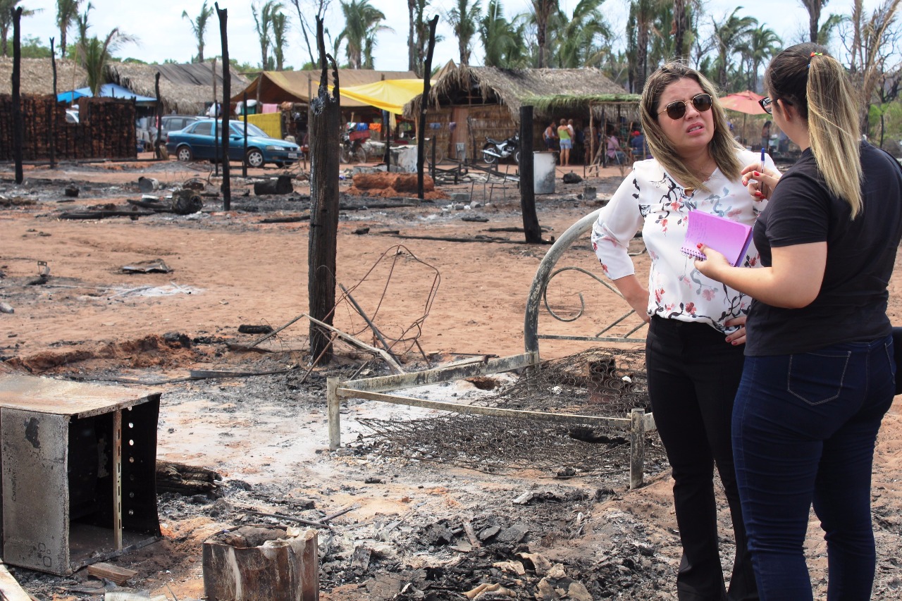 Duas pessoas de pé conversando, ao fundo estão os destroços provocados pelos incêndio que atingiu o assentamento 