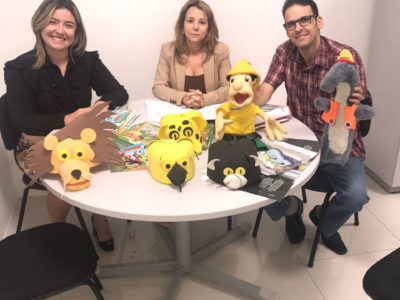Duas Promotoras de Justiça e um servidor do IBAMA sentados. Na frente deles, em uma mesa, há vários fantoches representando animais.