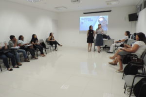 Pessoas sentadas em uma sala. Logo a frente dessas pessoas duas mulheres estão de pé falando com os que as assistem