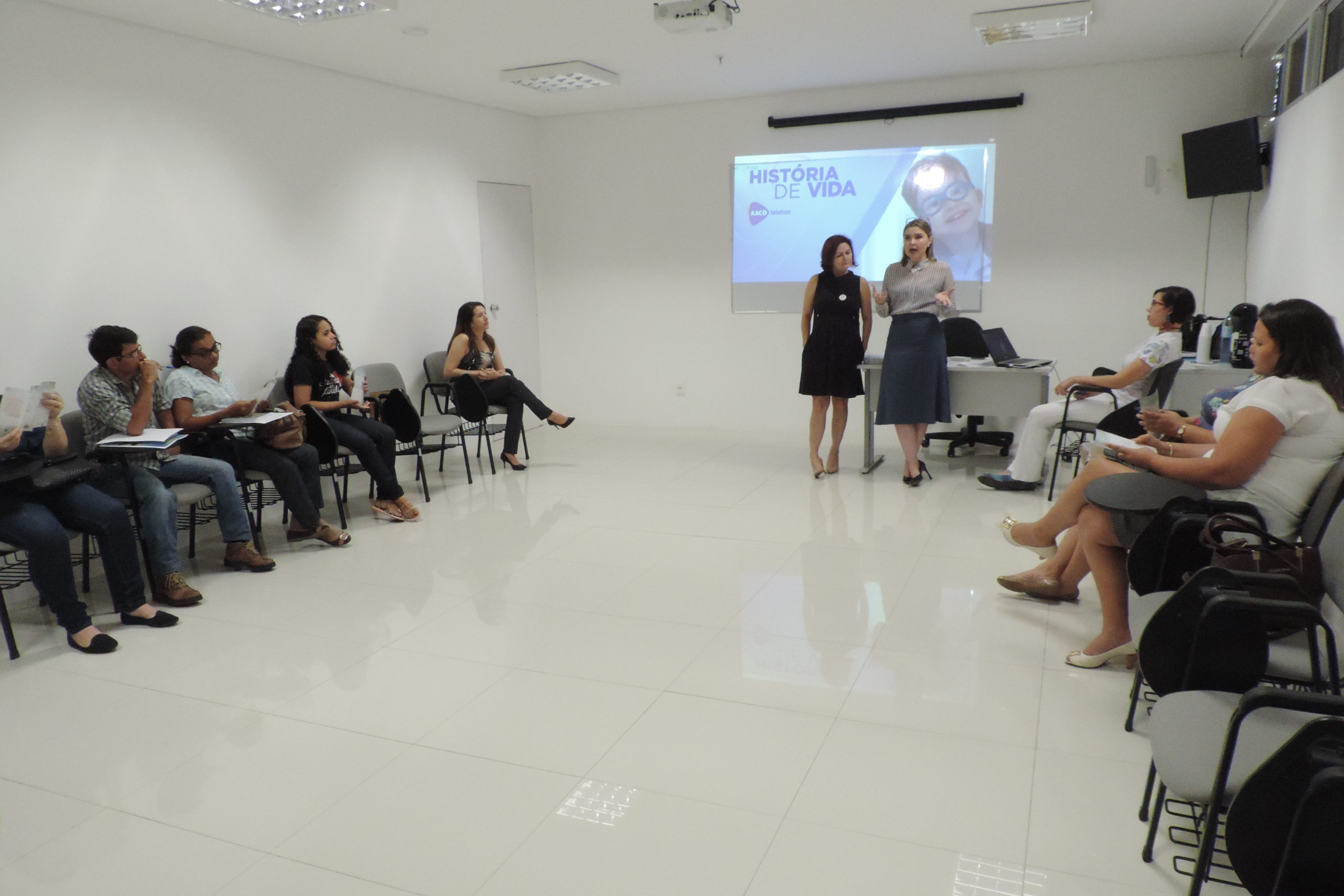 Pessoas sentadas em uma sala. Logo a frente dessas pessoas duas mulheres estão de pé falando com os que as assistem 